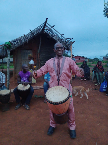 Man in traditional pink attire playing a drum while smiling, with people and dogs in the background near a thatched-roof hut.
