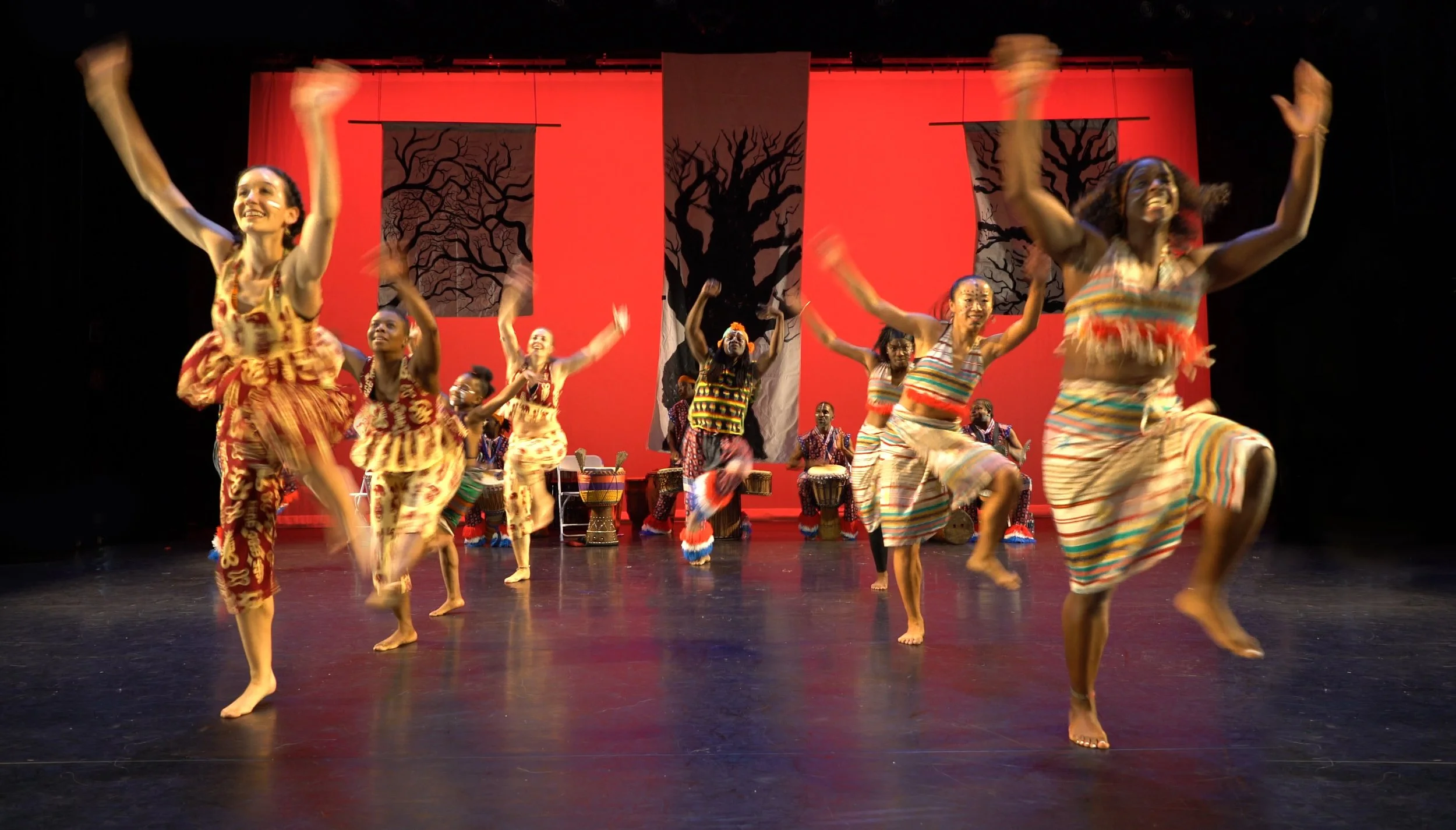 Group of dancers performing on stage, wearing colorful traditional African attire, with drums and a red background with black tree silhouettes.