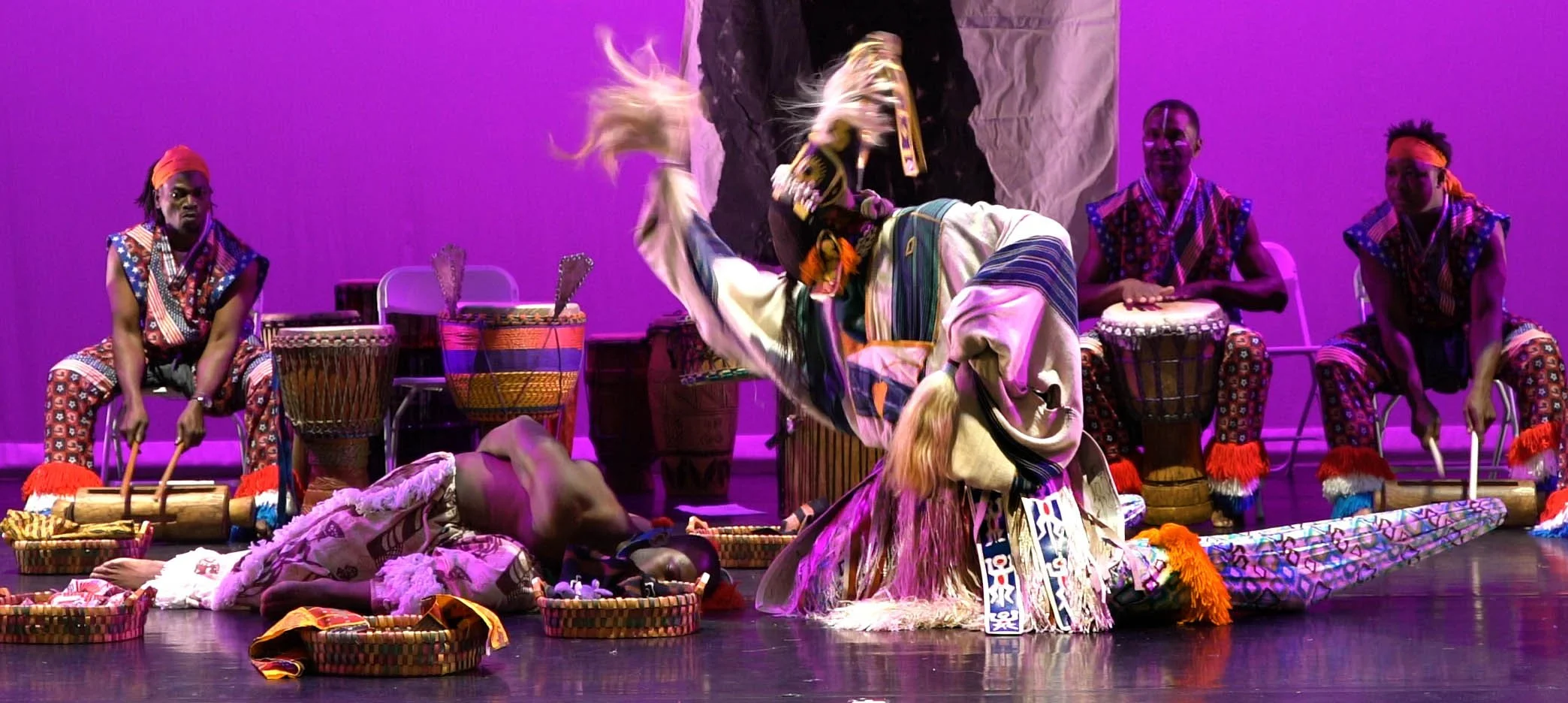 Performers in colorful traditional African costumes during a dance on stage, with drums and woven baskets around them, under purple stage lighting.