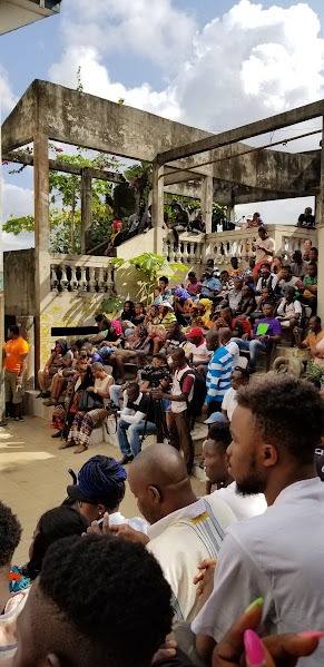 Crowd of people gathered outdoors on a concrete amphitheater with a modern structure, some sitting and others standing, under partly cloudy skies.