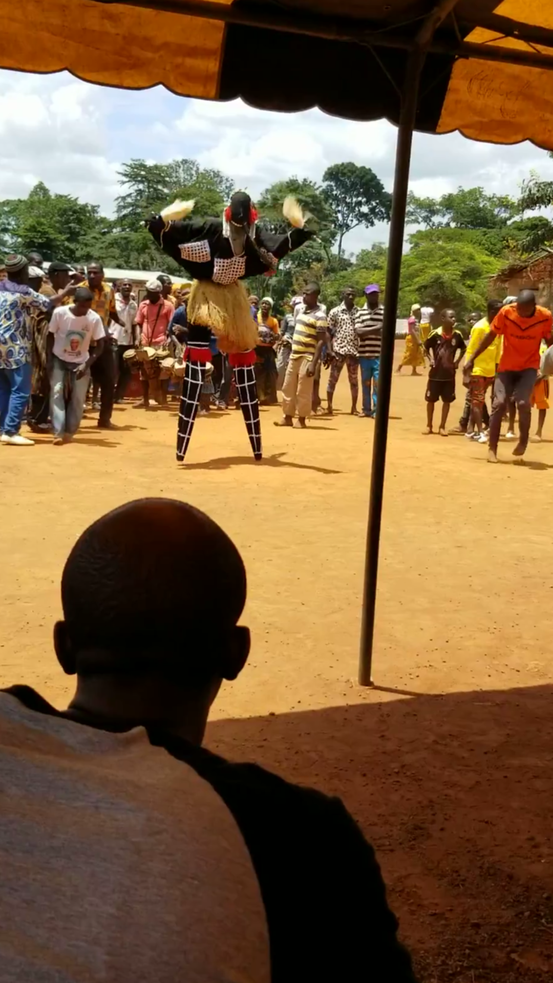 A performer on stilts in African traditional attire balancing a large puppet or figure on his head, surrounded by a crowd watching a cultural event outdoors.