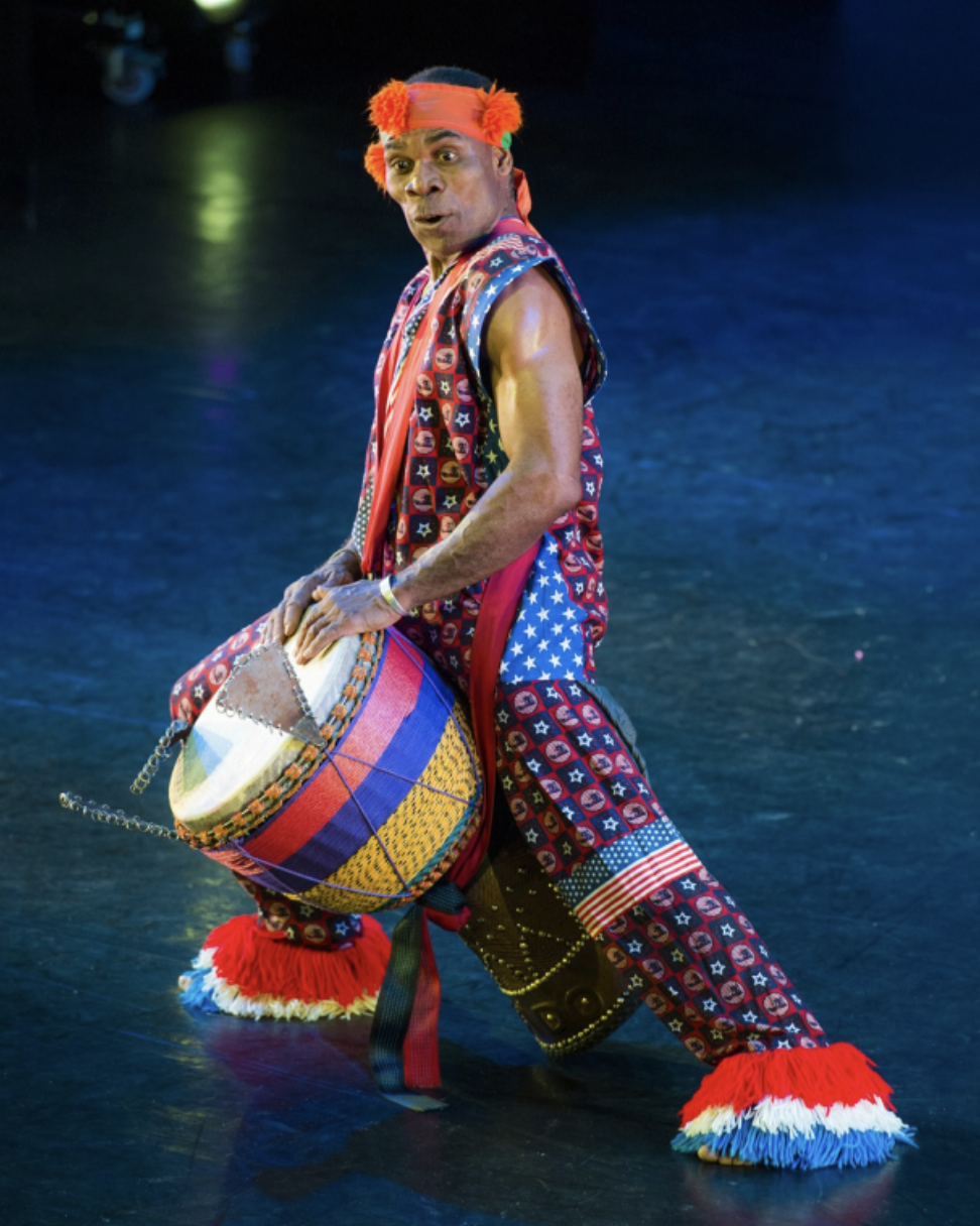 A man in colorful, patterned clothing performing with a decorated drum in a dark setting, with multi-colored stage lighting.