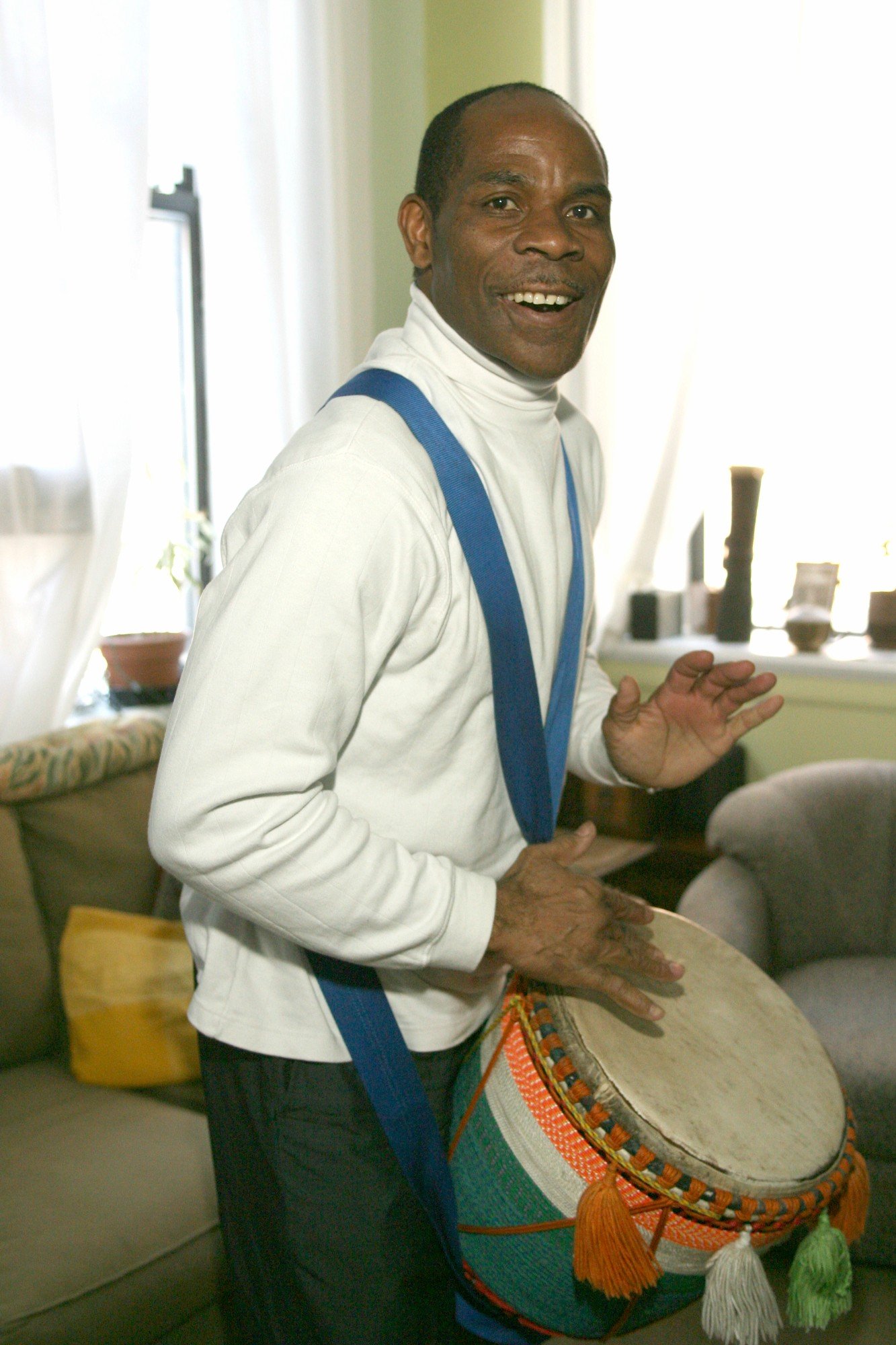 A man smiling and playing a drum inside a living room, wearing a white turtleneck and blue suspenders.