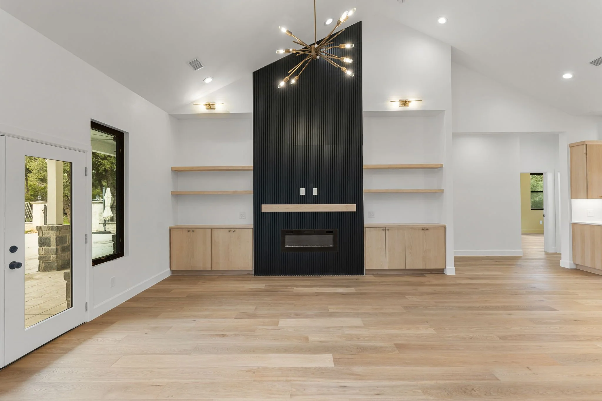 Empty living room with light wooden flooring, white walls, black accent wall with fireplace, wooden shelves, and a modern chandelier hanging from the ceiling.