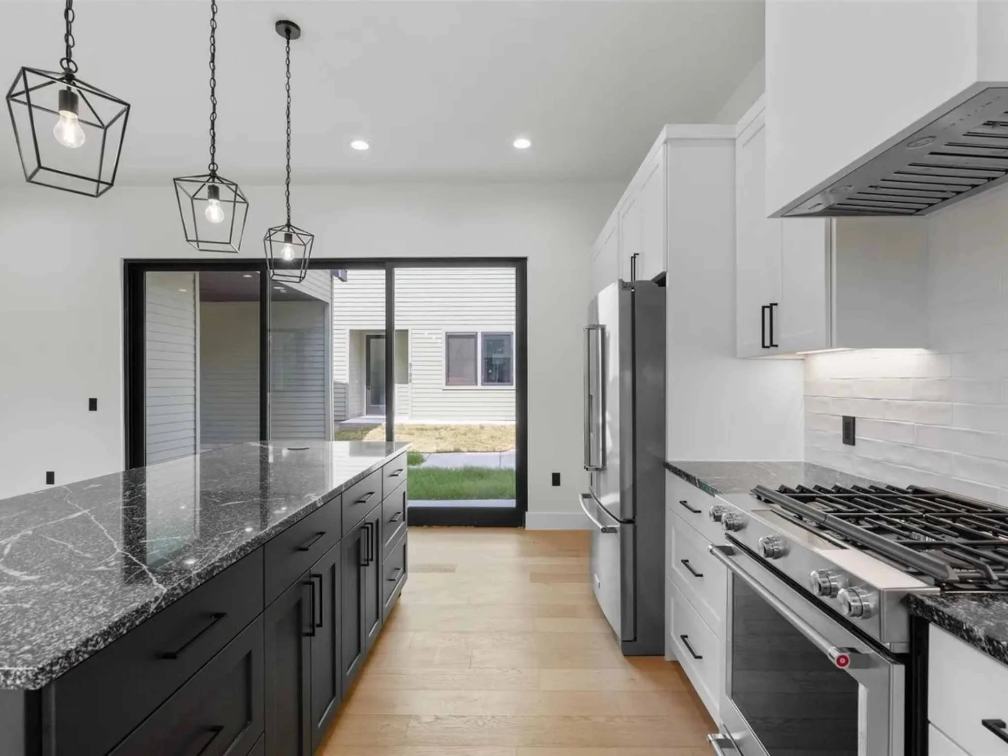 Modern kitchen with black island and white cabinets, stainless steel appliances, pendant lights with geometric metal frames, hardwood floors, and large sliding glass door leading to backyard.