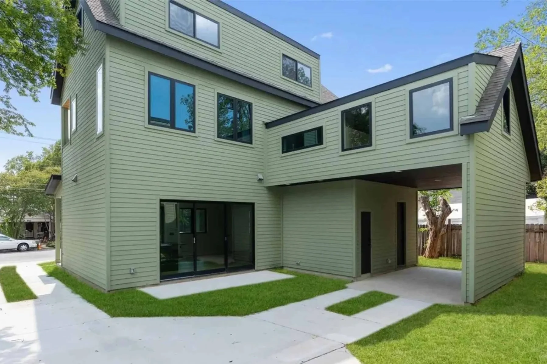 A modern multi-story house with green siding, multiple large windows, and a covered carport, surrounded by a green lawn and trees.