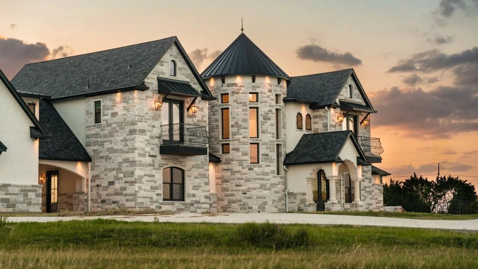 A large, elegant mansion with stone and stucco exterior walls, multiple gabled roofs, a round turret, and several balconies, set against a sunset sky.