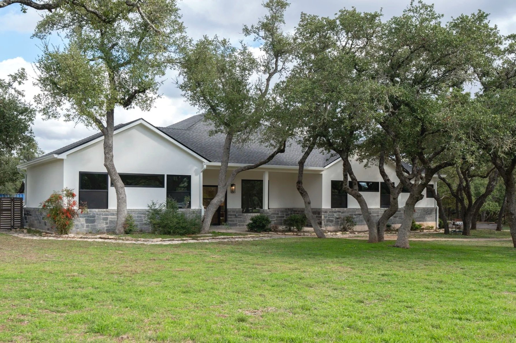 A single-story white house with a dark gray roof, multiple black-framed windows, a stone foundation, and a front porch, surrounded by a grassy yard with several trees and a cloudy sky overhead.