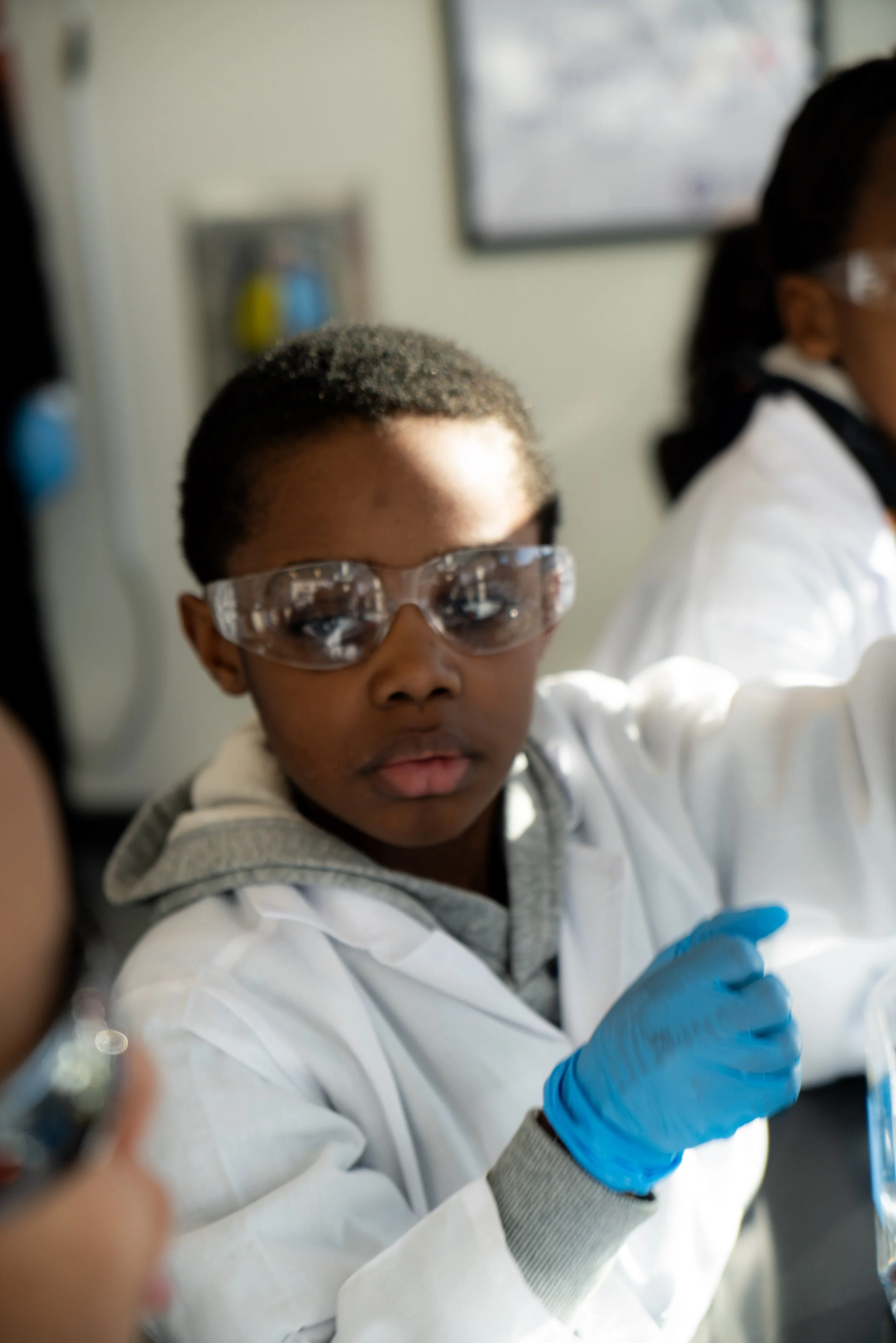 Young boy in safety goggles and a lab coat working on a science experiment in a classroom.
