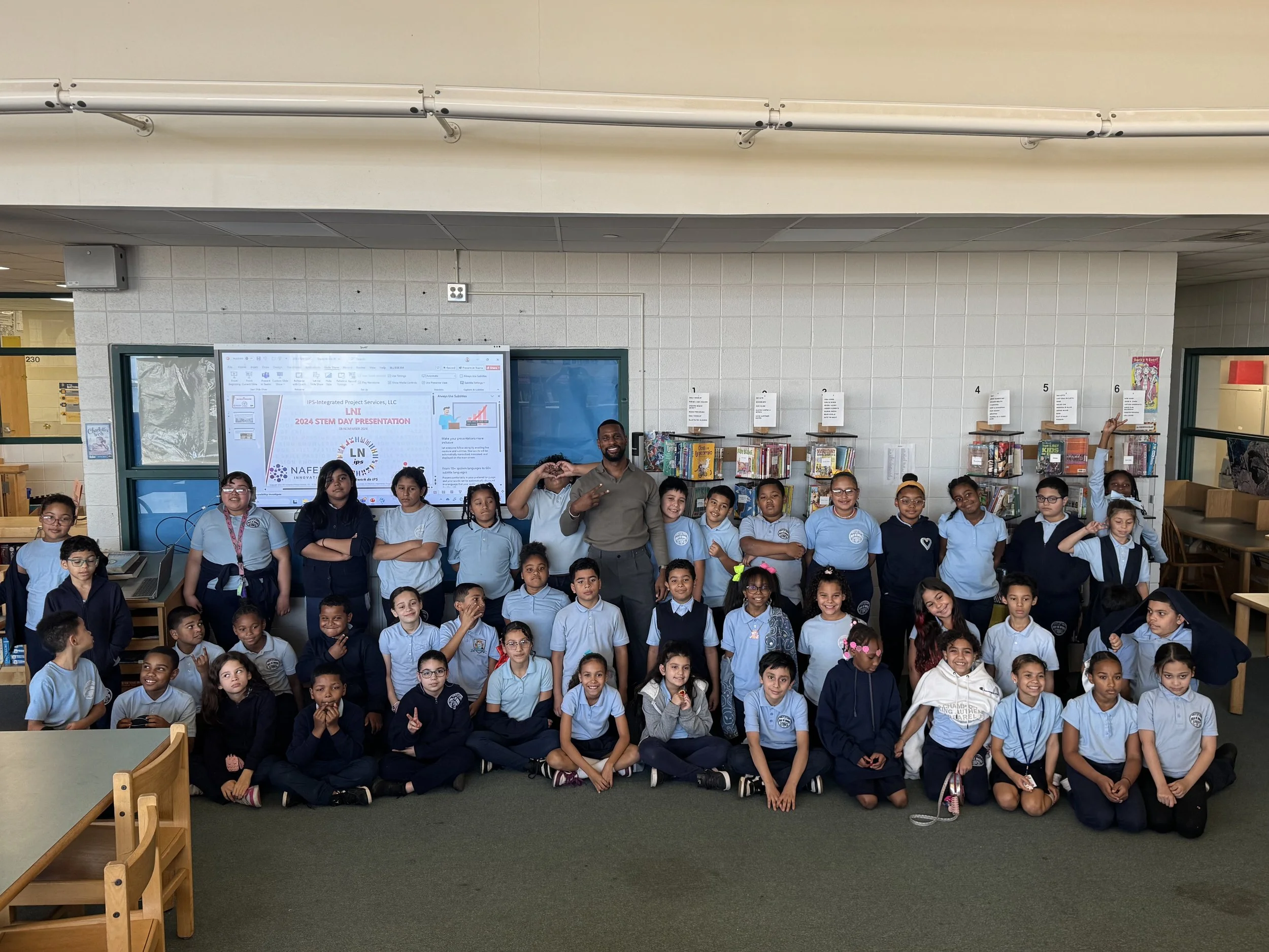 Group of elementary school students and a teacher posing for a photo in a library, with a presentation slide on a screen behind them that reads '2024 STEM DAY PRESENTATION'.