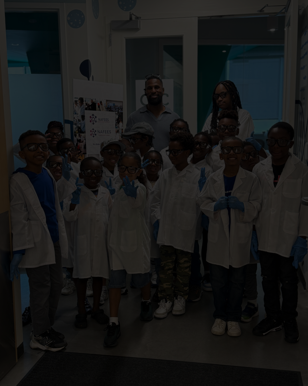 Group of children and two adults in a science lab, all wearing white lab coats and blue gloves, posing for a photo.