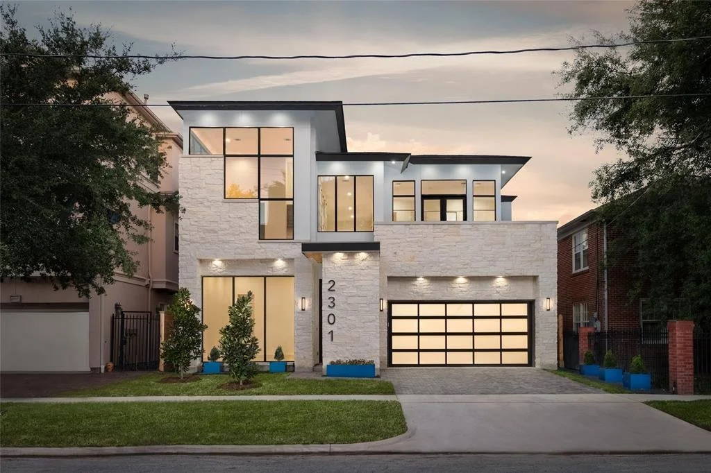 Modern multi-story house with large windows, a garage with horizontal panels, and outdoor lighting. The house has a white brick exterior and is surrounded by a neatly maintained front yard with small shrubs.