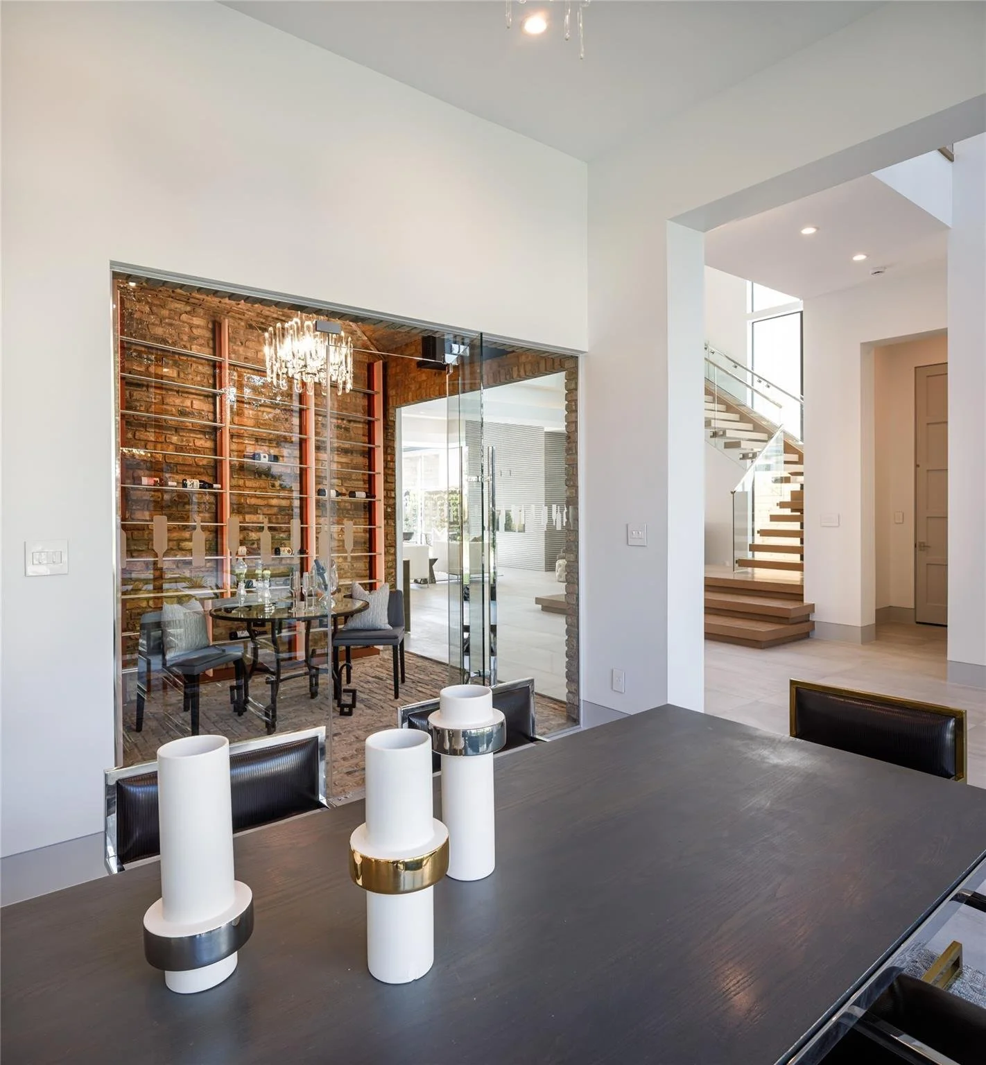 Modern dining room with a dark wood table, white and gold candles as centerpieces, and a glass wall revealing a brick-walled dining area with a chandelier and a chandelier.