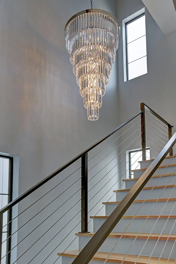 A modern staircase with wooden steps and metal railing in a well-lit interior, featuring a large, tiered crystal chandelier hanging from the ceiling and several windows providing natural light.