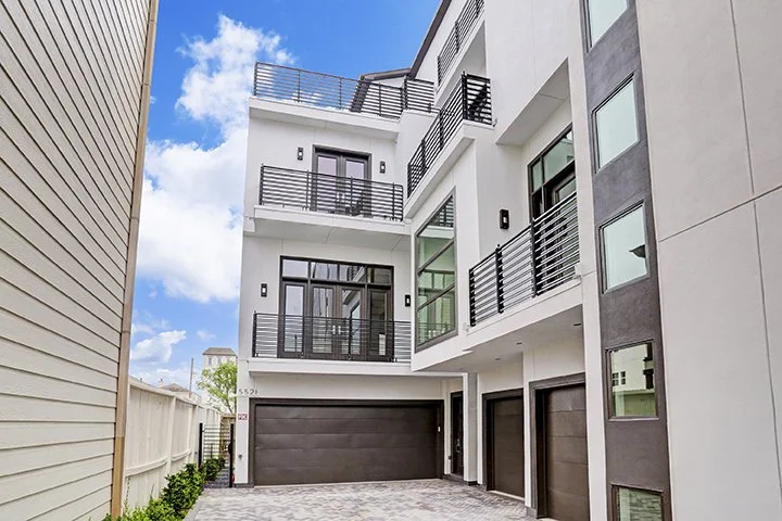Modern multi-story apartment building with balconies, black railings, glass balcony doors, and a gray garage door, with a clear blue sky in the background.