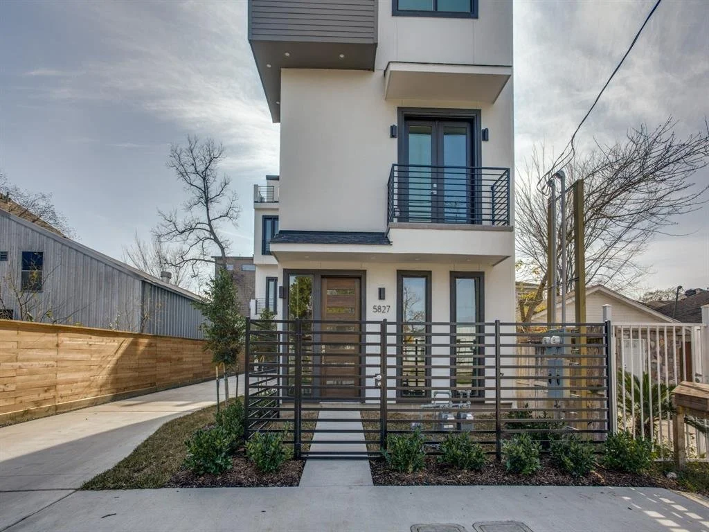 Modern multi-story white house with black railings and a small front yard with bushes, a sidewalk, and a driveway on the left. Bare trees and a cloudy sky are in the background.