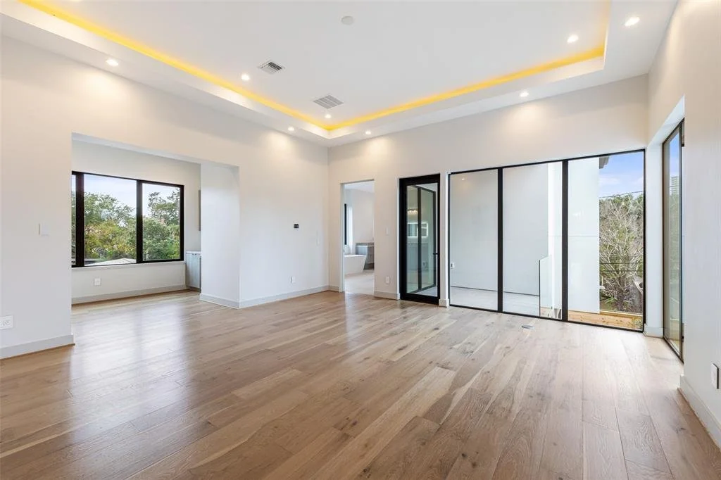Empty living room with hardwood floors, large windows, and sliding glass doors leading to a balcony, with recessed ceiling lights and yellow LED strip lighting.