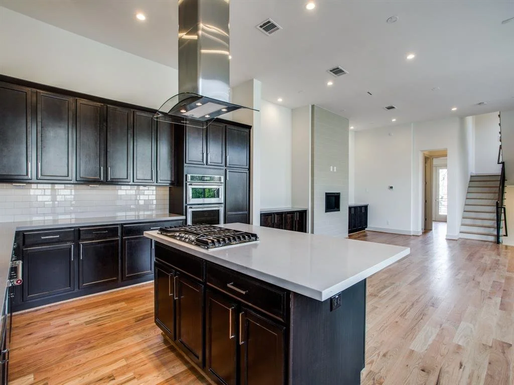 Modern kitchen with dark wood cabinets, white countertops, a built-in stove, an oven, and a range hood. Open living space with hardwood floors, white walls, and a staircase.