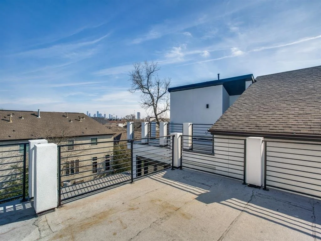 Rooftop view with a black railing, neighboring houses, a leafless tree, and a distant city skyline under a blue sky with wispy clouds.