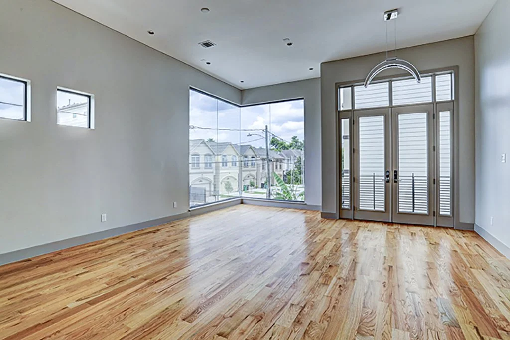 Empty living room with hardwood floors, large windows, and French doors leading outside, with small high windows near the ceiling.