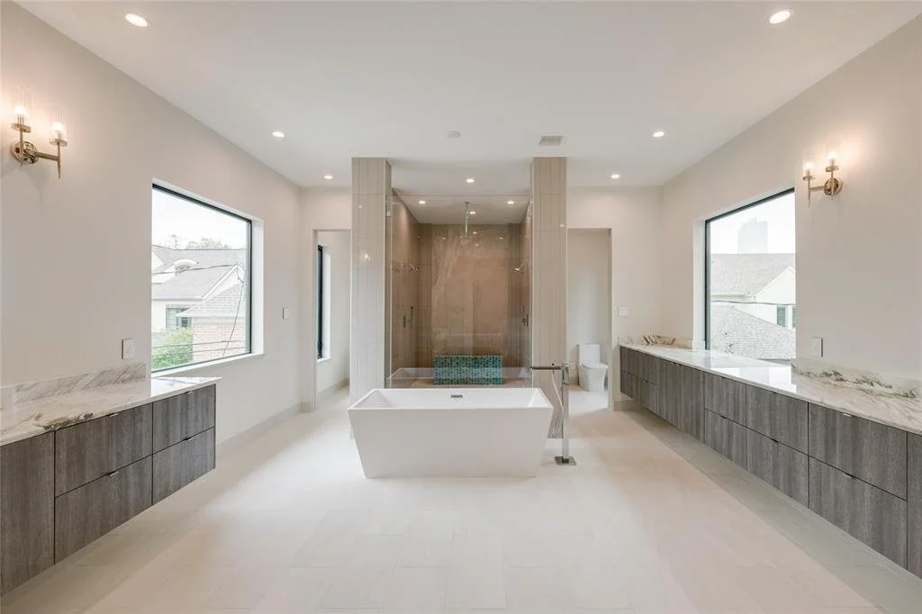 Modern bathroom with a freestanding bathtub, a glass-enclosed shower, twin vanities with marble countertops, and large windows with city views.