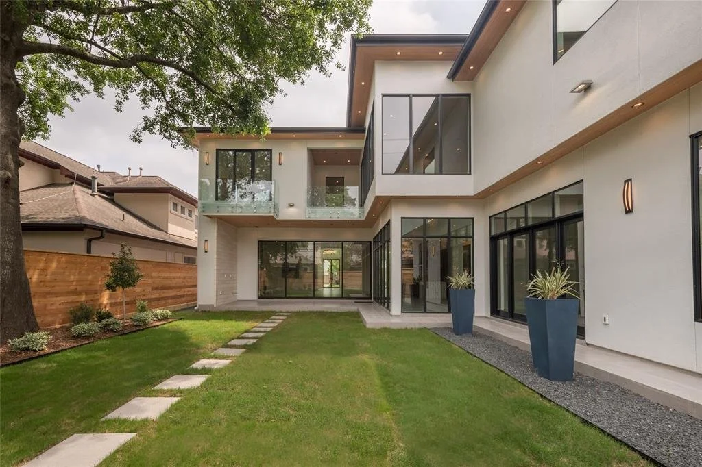 Modern two-story house with large glass windows and sliding doors, surrounded by a manicured lawn, potted plants, a tree, and a wooden fence.
