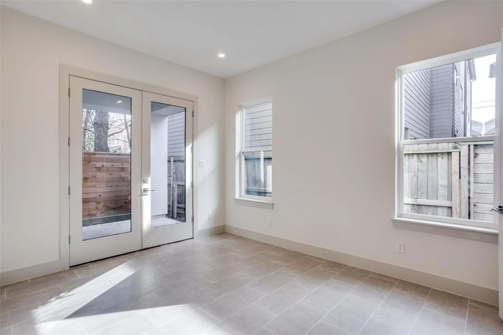 Empty room with white walls, beige tile floor, two windows, and glass door leading to a small outdoor area enclosed by wooden fencing.