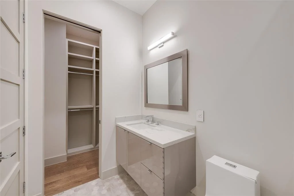 A bathroom with a white vanity, a mirror, a light fixture, a toilet, and a closet with shelves.
