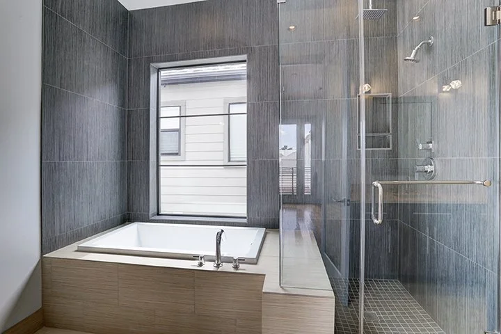 Modern bathroom with a bathtub, large window, and a glass-enclosed shower with dark gray tiles.
