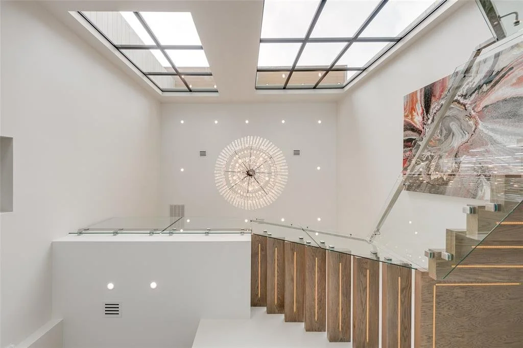 Modern staircase with glass railing, white walls, a large wall clock, and abstract artwork in a well-lit interior with a glass ceiling.