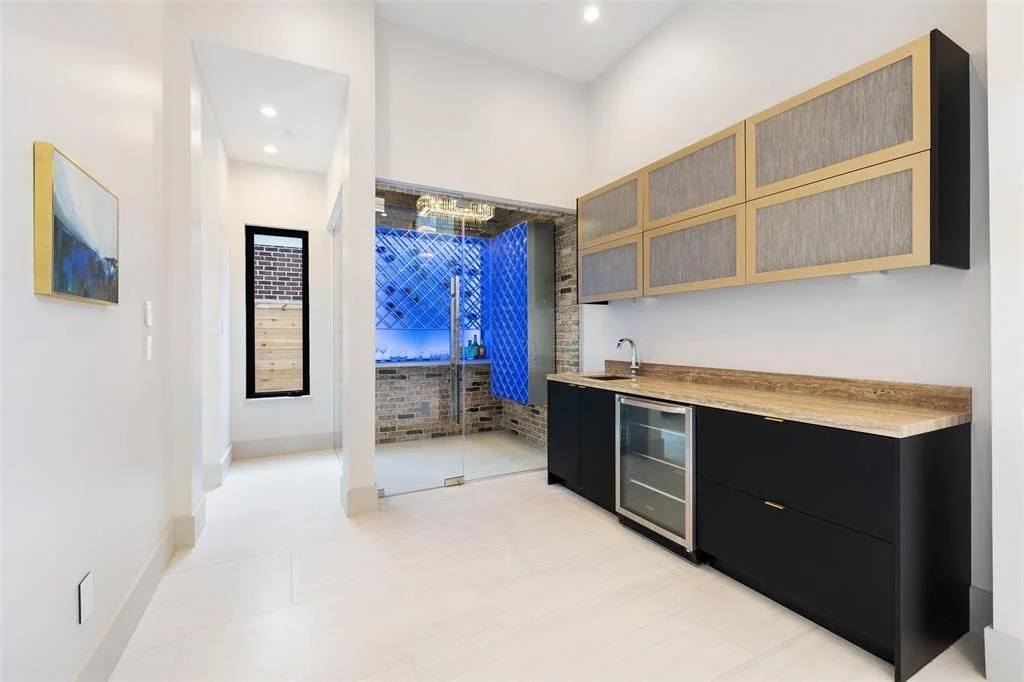 Modern kitchen with black lower cabinets, wooden countertop, wall-mounted cabinets with fabric panels, and a small wine fridge. There is a sliding glass door leading to an outdoor area with brick and wood elements, and a window to the left.