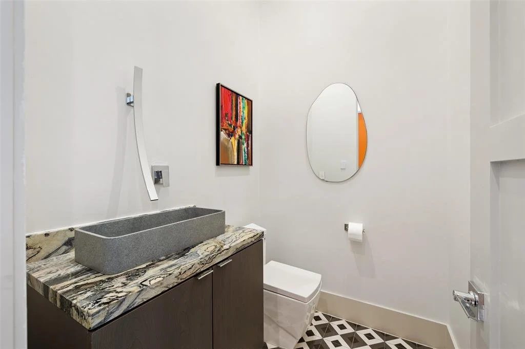Modern bathroom with a gray rectangular sink on a marble countertop, a wall-mounted faucet, a white toilet, an oval mirror, a piece of colorful abstract art, and patterned tile flooring.