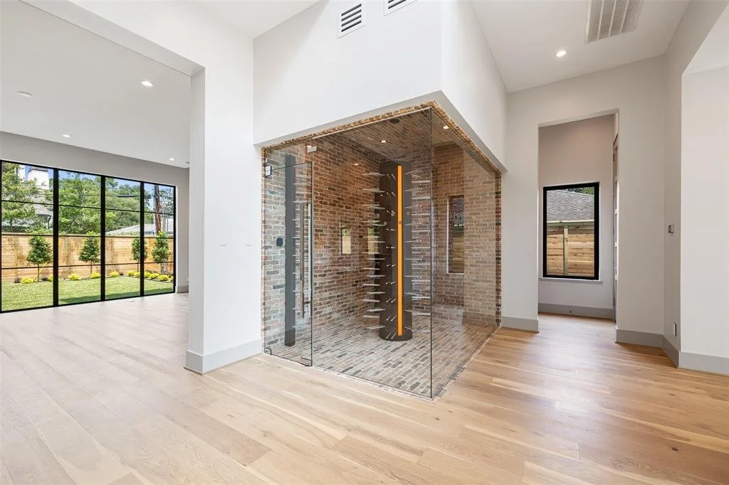 Modern interior with a glass sauna or steam room with exposed brick walls, surrounded by white walls and light wood flooring, with large windows showing a backyard.