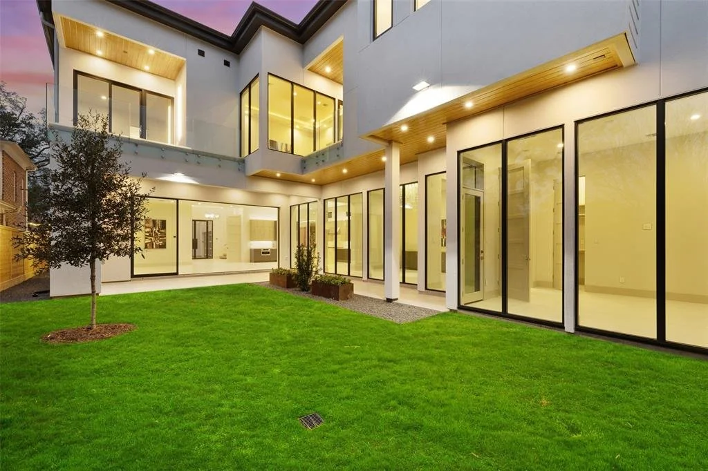 Modern multi-story house with large windows, a well-maintained green lawn, and exterior lighting at dusk.