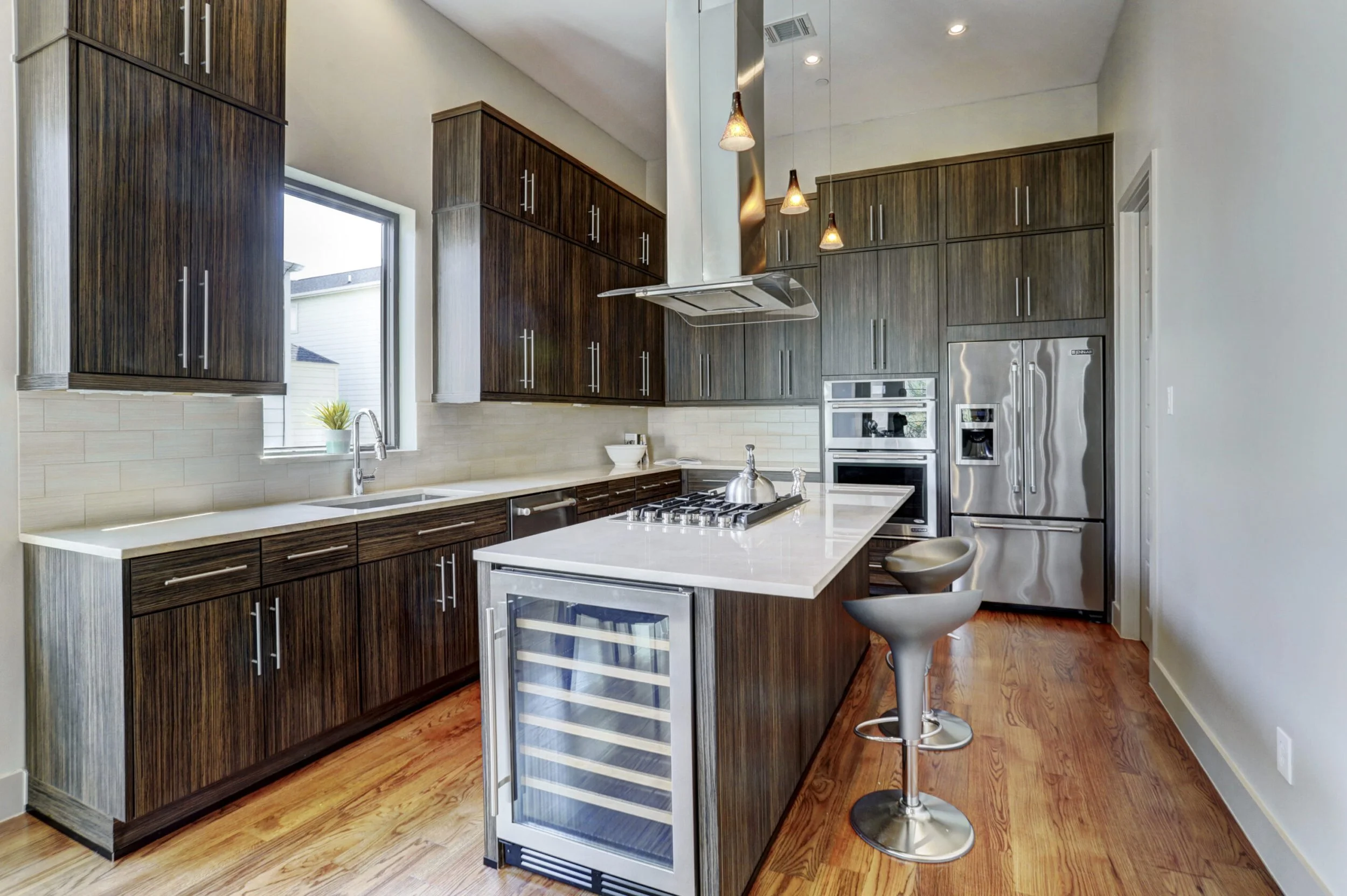 Modern kitchen with dark wood cabinets, white countertops, stainless steel appliances, a window above the sink, pendant lighting, a small bar with stools, and a wooden floor.
