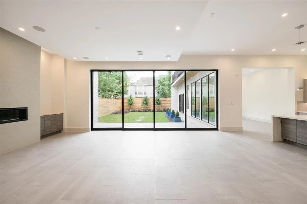 Empty modern living room with large glass sliding doors opening to a backyard with grass, trees, and a wooden fence.