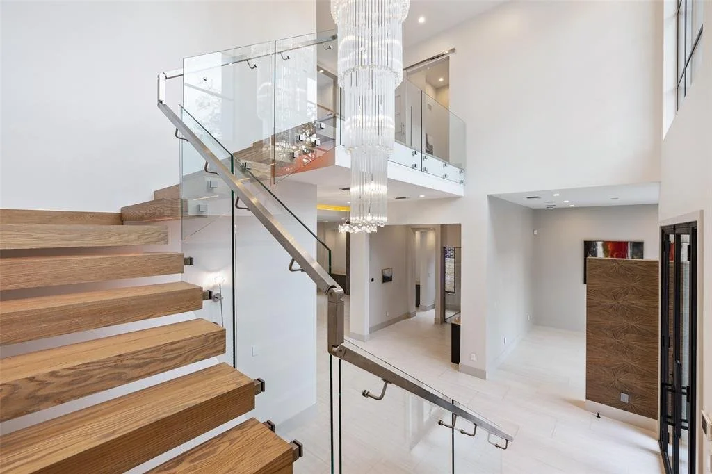 Modern interior staircase with wooden steps and glass railing leading to a spacious, open living area with white walls and high ceilings, featuring a large crystal chandelier.