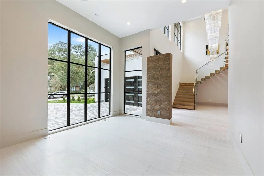 Interior view of a modern, bright living area with large floor-to-ceiling windows, a staircase with wooden steps and glass railing, and a decorative wall feature with a chandelier.