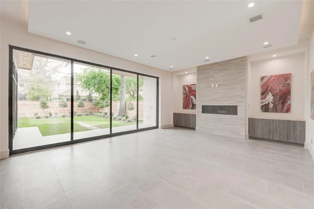 Empty living room with large glass doors leading to a backyard with trees and grass, modern fireplace feature wall, abstract art paintings, and neutral-colored walls and tiles.