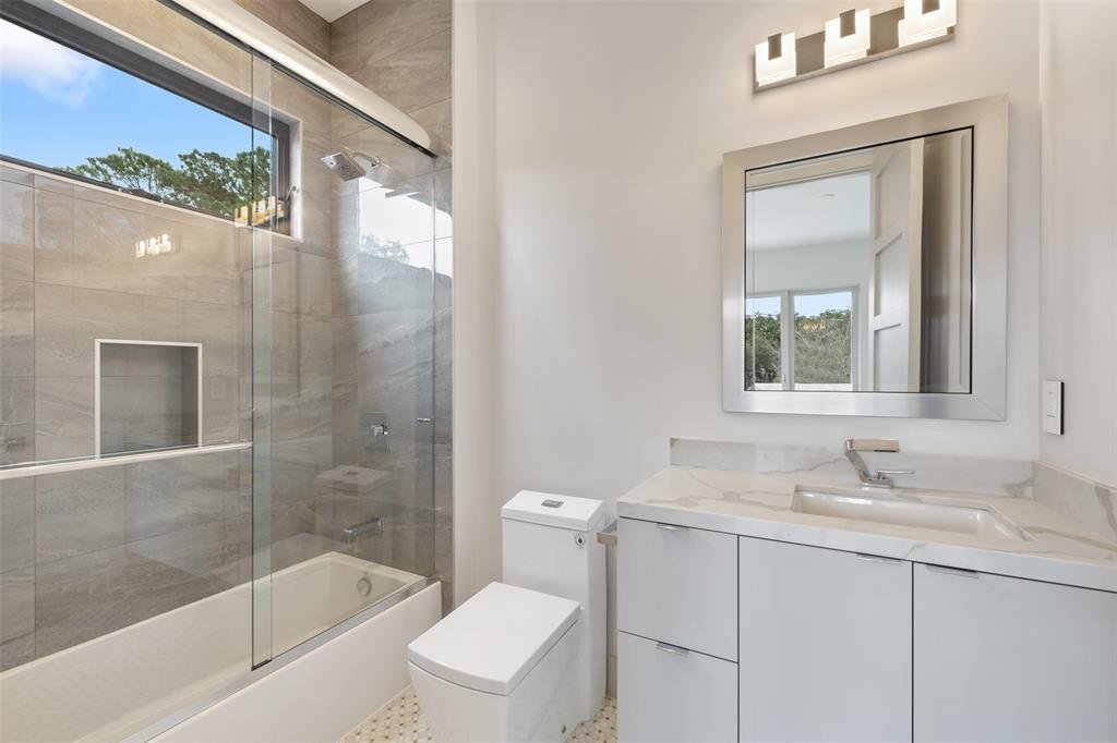 A modern bathroom featuring a glass-enclosed shower with a window, a white vanity with a marble countertop and mirror, a white toilet, and a row of contemporary light fixtures above the mirror.