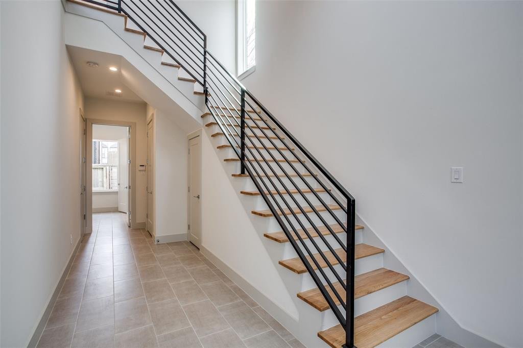 Interior view of a modern home hallway with a staircase featuring wooden steps and black metal railing, beige tiled floor, white walls, and natural light from a window.