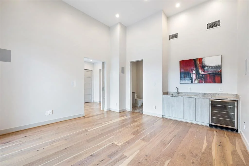 Empty room with hardwood floors, white walls, high ceiling with recessed lighting, a small kitchenette area with a marble countertop, a built-in wine cooler, and an abstract painting on the wall.