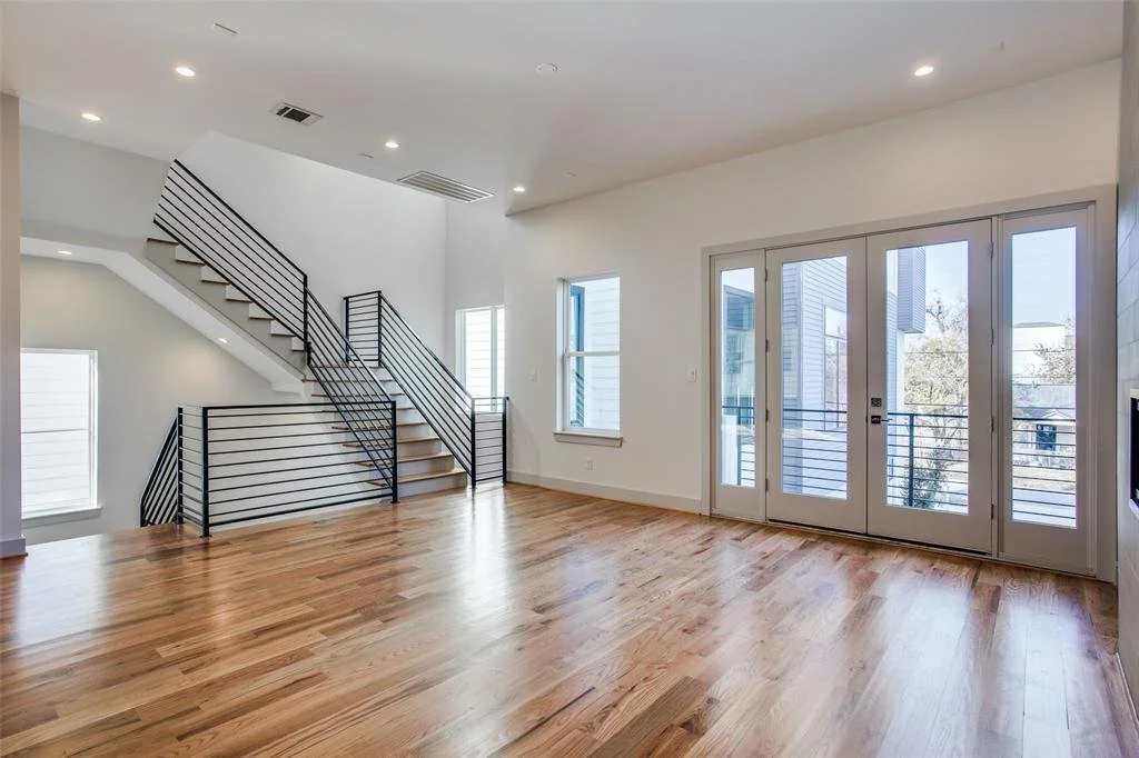 Empty modern living room with hardwood floors, large windows, and sliding glass doors leading to a balcony, featuring a staircase with black metal railings.