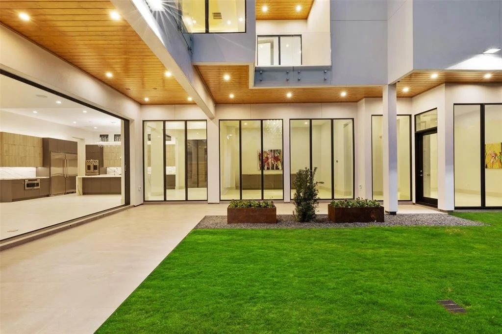 Modern house with large glass windows and doors, wooden ceiling accents, an indoor-outdoor design, a small garden with green grass and a couple of shrubs, and an open kitchen area.