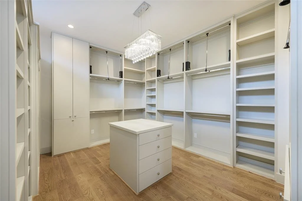 Empty walk-in closet with white built-in shelves, hanging rods, a small island in the center, and a chandelier overhead.