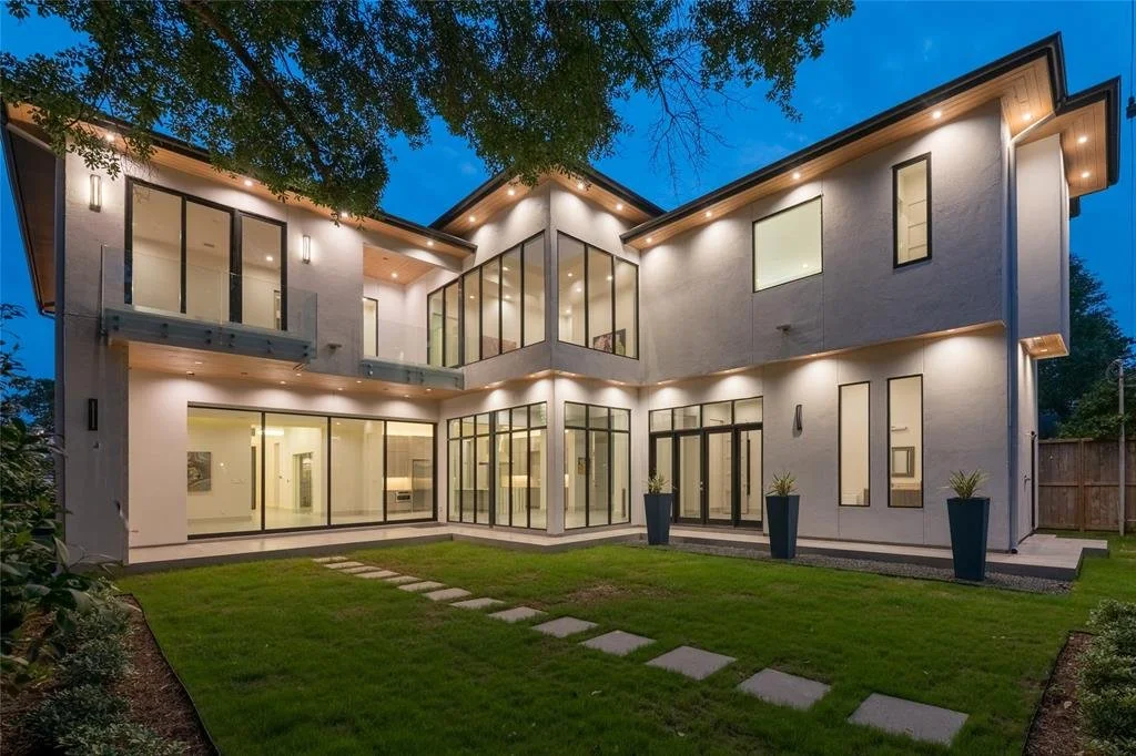 Modern multi-story house at dusk with large glass windows, white exterior walls, and landscaped front yard with stepping stones and tall planters with plants.