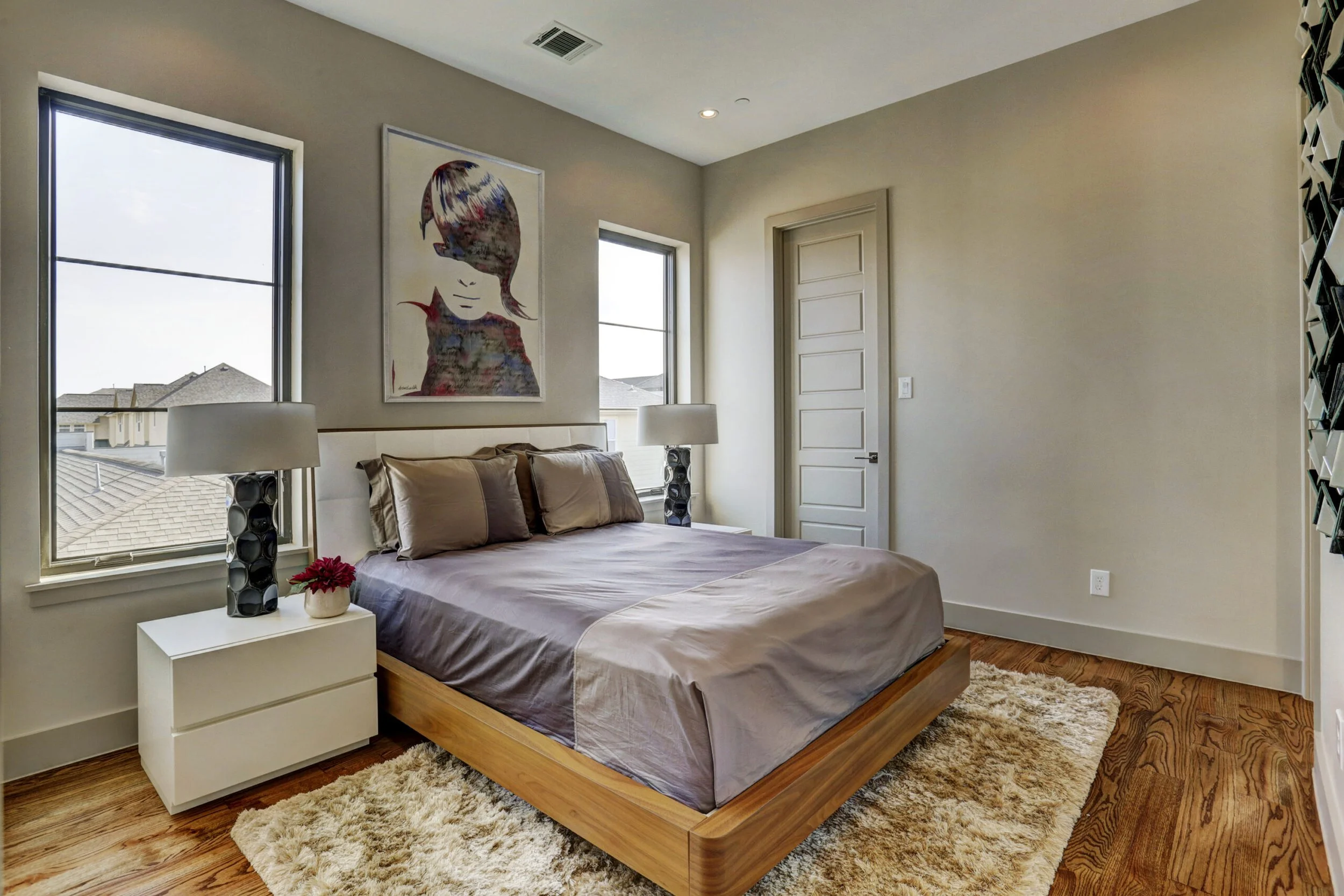 Bedroom with a wooden bed frame, beige bedding, twin nightstands with lamps, framed art of a girl with bangs, two windows, wooden door, and a beige rug.