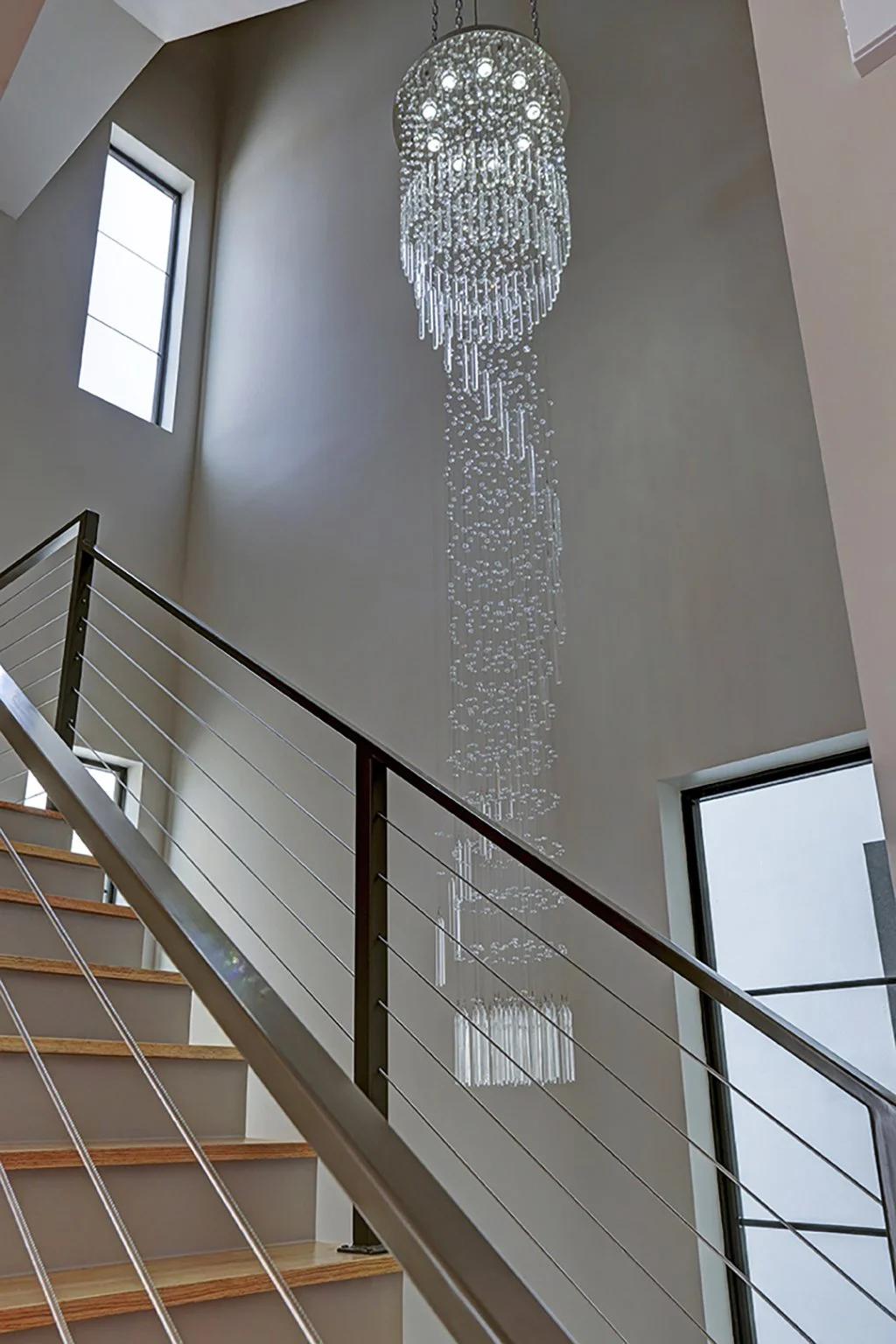 A modern staircase with wooden steps and metal railing, leading up to a high ceiling with a large, elaborate crystal chandelier hanging down.