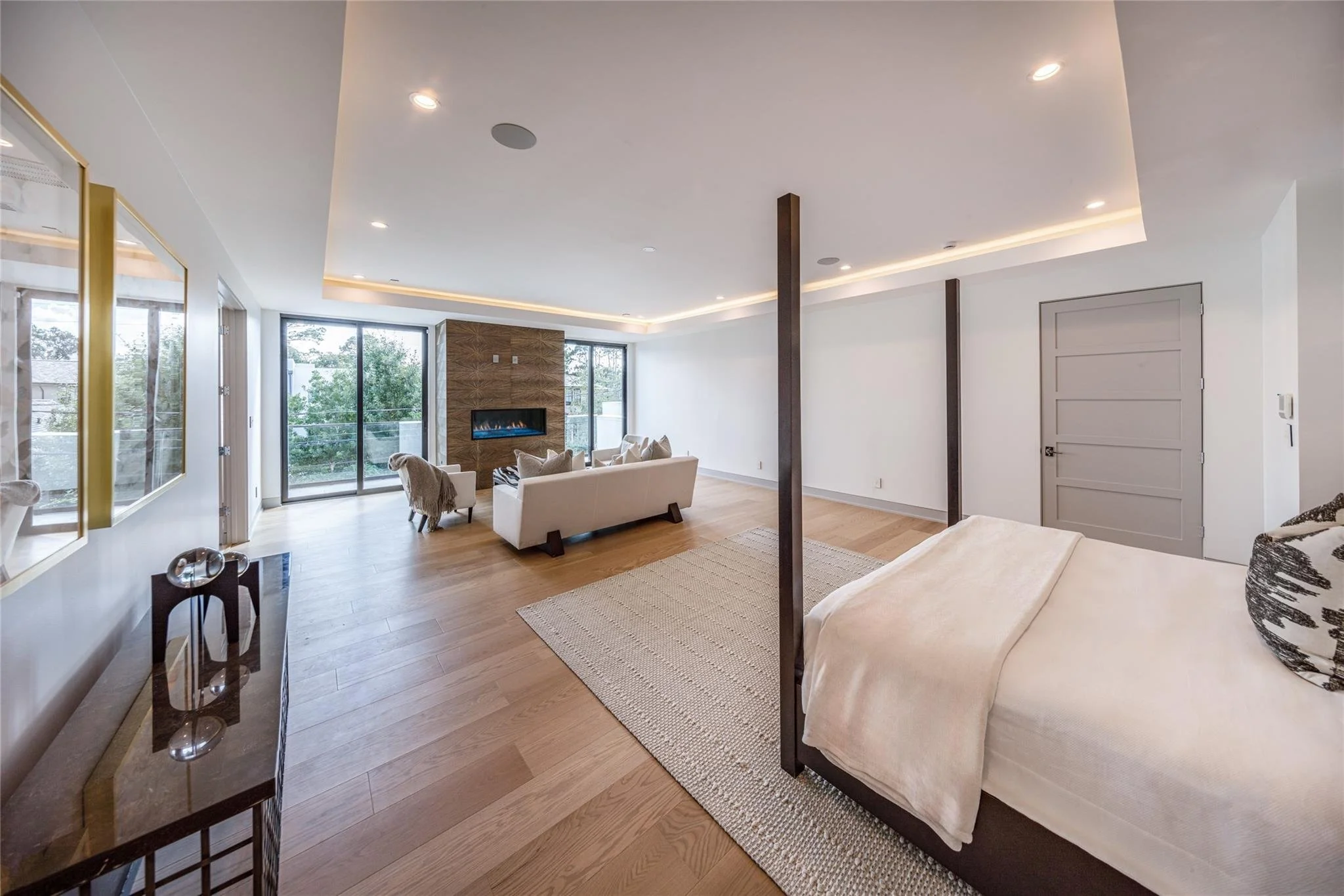 A spacious, modern living room with hardwood floors, a four-poster bed with white bedding, a white sofa, a stone fireplace, and large windows and sliding doors allowing natural light.