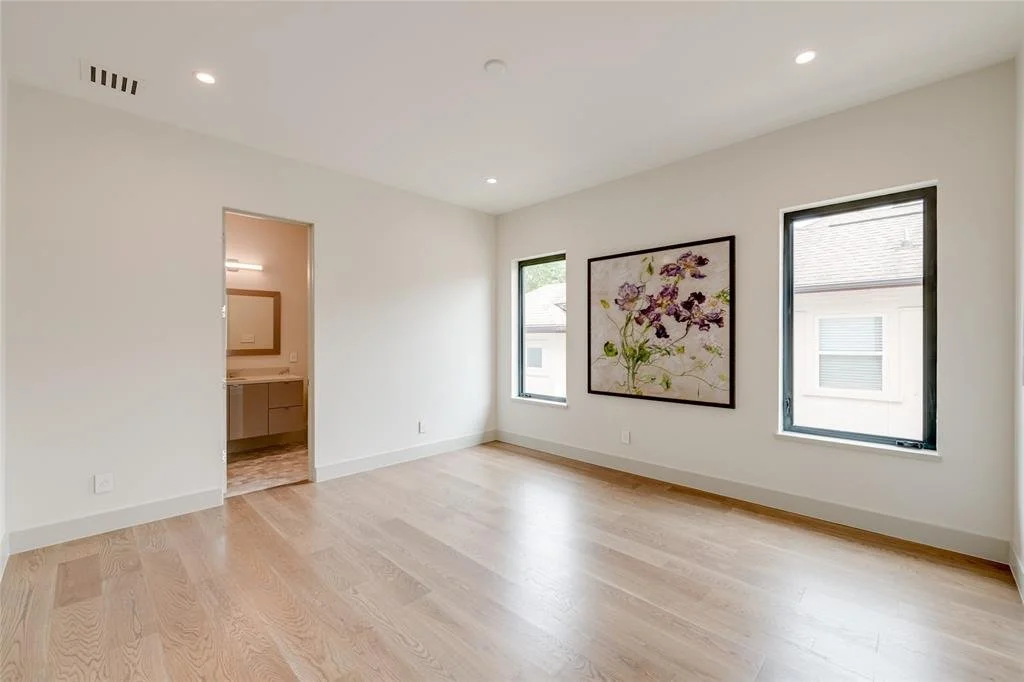 Empty room with white walls, light hardwood floors, two windows, and a large framed floral artwork on the wall.